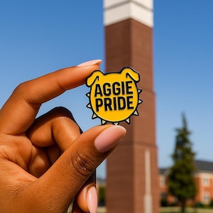 May include: A yellow and black enamel pin with the words "AGGIE PRIDE" in white lettering. The pin is in the shape of a dog's head. The background features a brick clock tower and a clear blue sky.