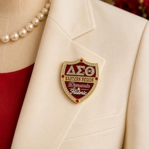 Op de afbeelding: Een schildvormige goudkleurige broche met een dieprood geëmailleerd midden, voorzien van de Griekse letters Delta Sigma Theta in heldere strass-steentjes, de tekst EASTERN REGION en de woorden Dynamic & Historic, omrand door kleine steentjes.
