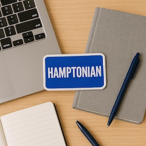 以下が含まれることがあります： 「HAMPTONIAN」の文字が入った青と白のパッチが、木製の机の上に置かれています。ノートパソコン、グレーのノート、2本の青いペンも一緒に写っています。パッチには白い縁取りがあります。