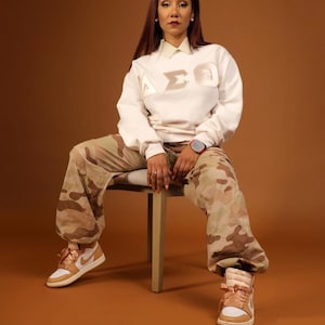 Puede incluir: Una mujer con una sudadera blanca con las letras "AΣΘ" en color crema en la parte delantera, pantalones de camuflaje y zapatillas altas de color marrón claro. Está sentada en una silla de madera sobre un fondo marrón.