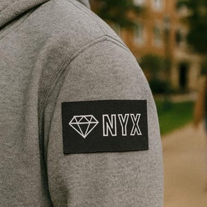 以下が含まれることがあります： ダイヤモンドのグラフィックと白い「NYX」の文字が入った黒いパッチが付いたグレーのパーカー。 パーカーは柔らかい素材でできており、ドローストリングフードが付いています。 画像は屋外で撮影されました。