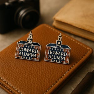 May include: Two Howard University alumni pins. Each pin is shaped like a building with a silver outline, a blue roof, and a red brick facade. One pin reads "HOWARD ALUMNA" and the other reads "HOWARD ALUMNI". The pins are on a brown leather surface.