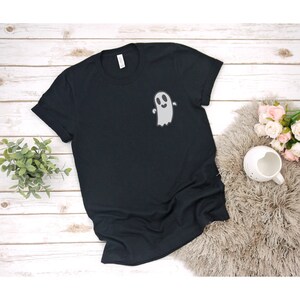 以下が含まれることがあります： 前面に白い幽霊のグラフィックがプリントされた黒いTシャツ。幽霊は笑顔で腕を伸ばしています。
