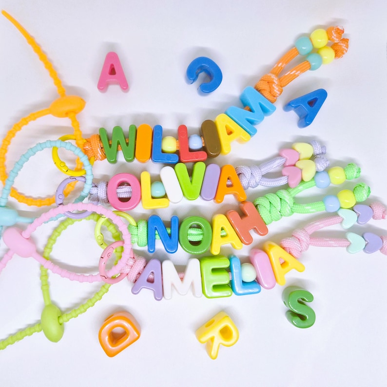 May include: Four personalized keychains made of colorful plastic letter beads on nylon cords, spelling the names WILLIAM, OLIVIA, NOAH, and AMELIA. The keychains feature various pastel and bright colored beads, including heart shapes and loops.