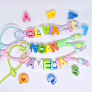 May include: Three personalized keychains featuring colorful plastic letter beads spelling OLIVIA, NOAH, and AMELIA. Each keychain includes a nylon cord, a circular ring, and additional decorative beads in shapes like hearts and spheres.