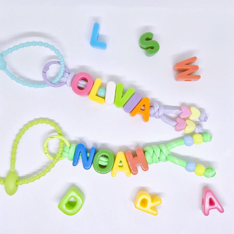 May include: Two personalized keychains featuring colorful plastic letter beads spelling OLIVIA and NOAH. The keychains are attached to light blue and lime green silicone beaded loops with additional heart-shaped beads and loose alphabet letters.