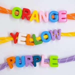 May include: Three strands of cord in orange, yellow, and purple, each threaded with colorful plastic alphabet beads. The top strand spells ORANGE, the middle strand spells YELLOW, and the bottom strand spells PURPLE using various letter colors.