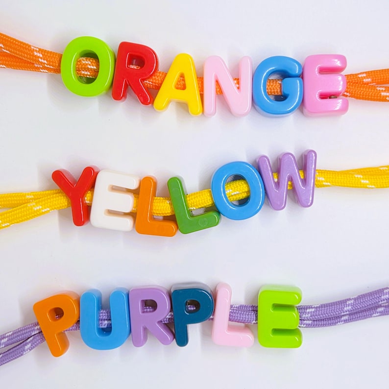 May include: Three strands of cord in orange, yellow, and purple, each threaded with colorful plastic alphabet beads. The top strand spells ORANGE, the middle strand spells YELLOW, and the bottom strand spells PURPLE using various letter colors.
