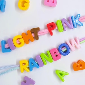 May include: Two strands of light pink and light blue nylon cord are threaded with colorful plastic alphabet beads. The top strand spells LIGHT PINK in multi-colored letters, while the bottom strand spells RAINBOW in various bright plastic letters.