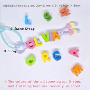 May include: A personalized keychain featuring colorful alphabet beads spelling OLIVIA on a lavender cord, attached to a light blue silicone strap and a purple O-ring. Alphabet beads measure 20-21mm x 14-18mm x 8mm. Includes loose letter beads.