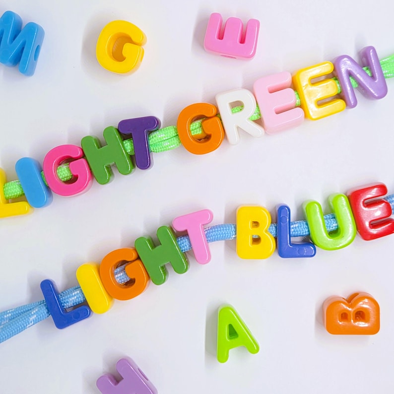 May include: Two strands of neon-colored cord threaded with multi-colored plastic alphabet beads. The top strand spells LIGHT GREEN and the bottom strand spells LIGHT BLUE. Additional loose plastic letter beads in various colors are scattered around.