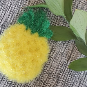 May include: A yellow and green crocheted pineapple on a grey textured surface.