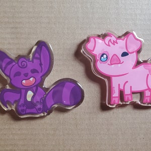 Puede incluir: Dos colgantes de acrílico de dibujos animados. Uno es un murciélago morado con una cola a rayas y una expresión feliz. El otro es un cerdo rosa guiñando un ojo, con un ojo azul y una mancha roja en la espalda. Ambos colgantes tienen un borde transparente.