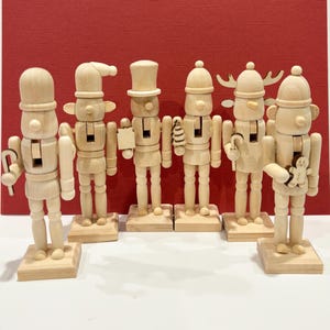 May include: Six wooden nutcrackers with different hats and accessories, standing on individual wooden bases. The unpainted figures are set against a red backdrop. Each nutcracker holds a small item, such as a tree or a gingerbread man.