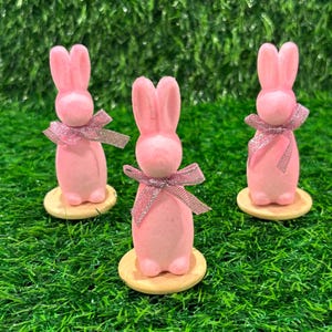 Pode incluir: Três figuras de coelhos cor-de-rosa com laços de glitter prateado, cada uma sobre uma pequena base redonda de madeira. Os coelhos têm orelhas eretas e estão sobre um fundo de relva artificial verde, criando um tema de Páscoa.