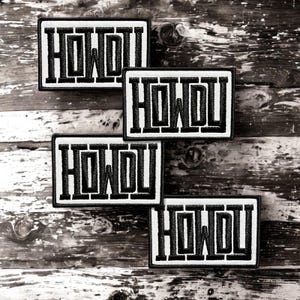 May include: Four rectangular embroidered patches featuring the word HOWDY in bold black block letters on a white background. Each patch has a thick black border and a textured fabric appearance, arranged in a scattered overlapping pattern.