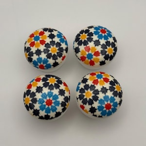 May include: Four decorative cabinet knobs with a white base and a colourful starburst pattern. The pattern features navy, red, yellow, and blue star shapes. These knobs are a decorative accent for furniture.