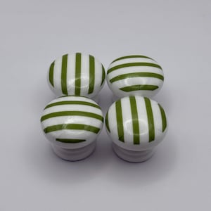 May include: Four white cabinet knobs featuring a green striped design. Each knob has a rounded top with a green striped pattern and a short, cylindrical base. The knobs are arranged in a square formation. The green stripes are evenly spaced.