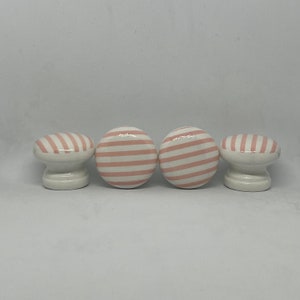 May include: Four white ceramic cabinet knobs with a pink and white striped pattern. Two knobs are round, and two are oval-shaped. The knobs are suitable for drawers, cabinets, or furniture. The knobs are on a white background.