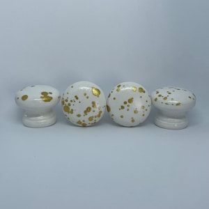 May include: Four white round knobs with gold splatter paint. The knobs are approximately 2.5 cm in diameter.