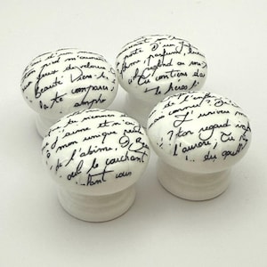 May include: Four white ceramic cabinet knobs in a mushroom shape. Each knob features handwritten black script, possibly a poem or quote. The knobs are set against a white backdrop.