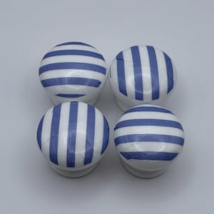 May include: Four white cabinet knobs with blue vertical stripes. The knobs are round with a small, white base. The blue stripes are evenly spaced across the surface of the knobs. The knobs are arranged in a square.