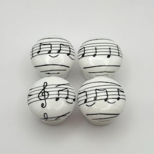 May include: Four white ceramic cabinet knobs featuring black musical notation. Each spherical knob is decorated with a musical stave and notes. These knobs would be suitable for a music-themed room or furniture.