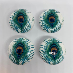 May include: Four round white ceramic knobs featuring a peacock feather design. The feathers are blue, green and gold with a blue eye in the centre.