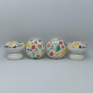 May include: Four white ceramic knobs with a floral pattern in shades of yellow, green, pink, and purple. The knobs have a rounded top and a base.