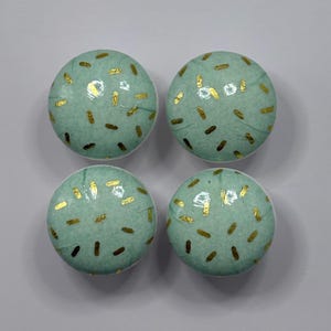 May include: Four round, light teal cabinet knobs with a speckled gold pattern. The knobs are arranged in a square formation, each with a smooth, glossy finish. These decorative knobs are ideal for adding a touch of colour and style to furniture.