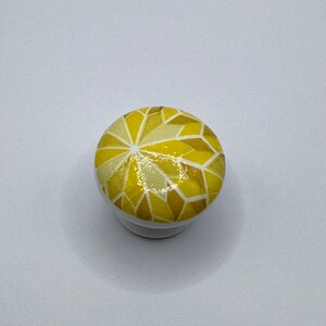 Peut inclure: Bouton de meuble rond blanc avec un motif g&eacute;om&eacute;trique dans les tons de jaune et d'or. La surface sup&eacute;rieure du bouton pr&eacute;sente un motif en forme de mosa&iuml;que, ajoutant une touche d&eacute;corative aux meubles ou aux armoires. Petit &eacute;l&eacute;ment de quincaillerie fonctionnel.