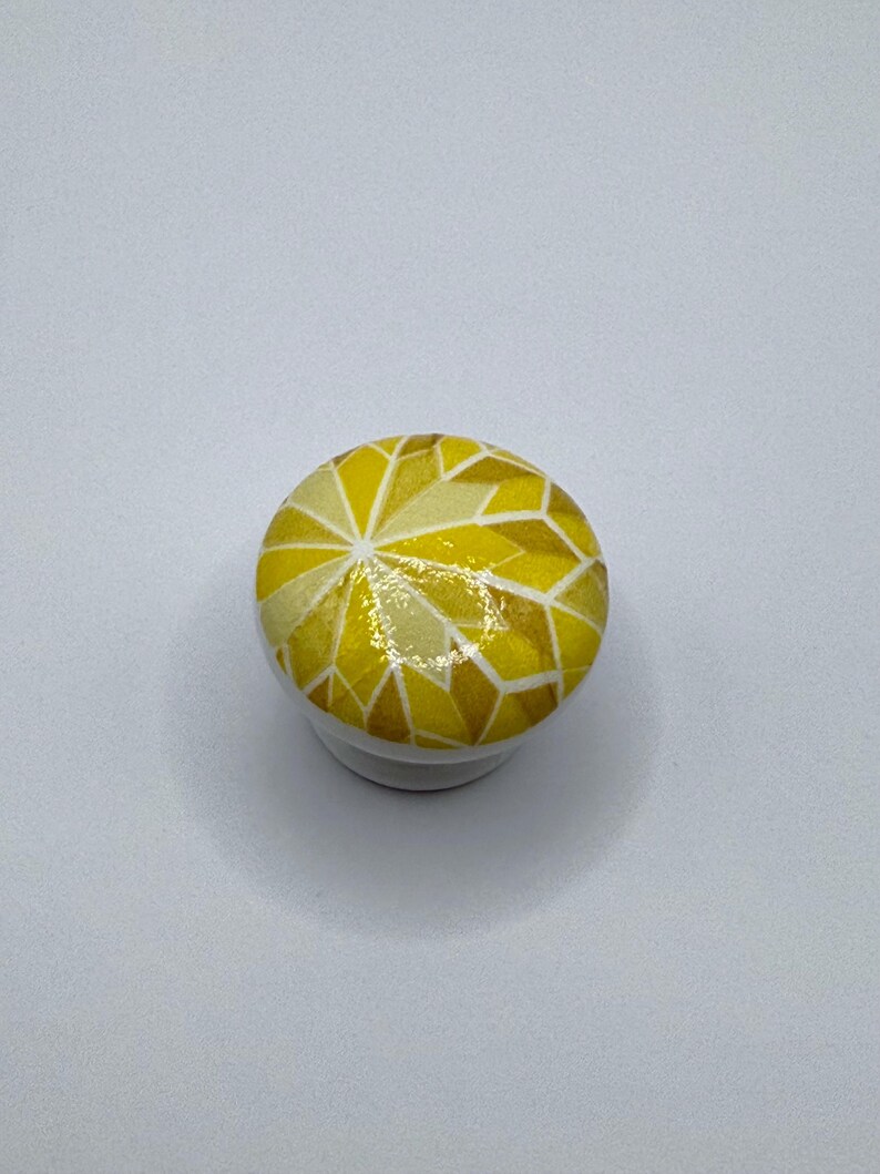 Peut inclure: Bouton de meuble rond blanc avec un motif g&eacute;om&eacute;trique dans les tons de jaune et d'or. La surface sup&eacute;rieure du bouton pr&eacute;sente un motif en forme de mosa&iuml;que, ajoutant une touche d&eacute;corative aux meubles ou aux armoires. Petit &eacute;l&eacute;ment de quincaillerie fonctionnel.