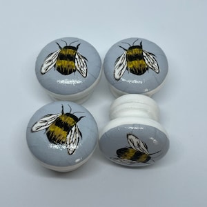 May include: Four round wooden knobs with a grey background and a painted bee design. The bees are yellow and black with brown stripes and white wings.