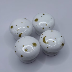 May include: Four white ceramic cabinet knobs with gold celestial designs. Each knob features gold stars and crescent moons. The knobs have a rounded shape and a small, white base.