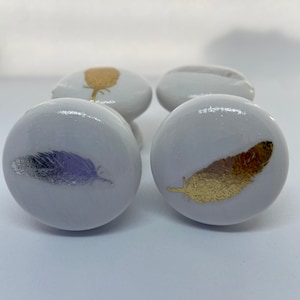 May include: Four white ceramic cabinet knobs with feather designs. Two knobs feature gold feathers, one has a silver feather, and another has a gold design. The knobs are round and have a glossy finish, suitable for furniture upgrades.