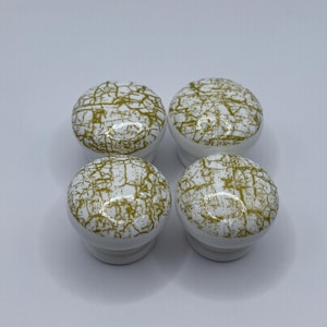 May include: Four white ceramic cabinet knobs with a gold crackle pattern. The knobs have a rounded shape and a smooth, glossy finish. These decorative knobs are ideal for adding a touch of elegance to furniture or cabinetry.