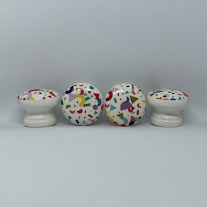 May include: Four white ceramic cabinet knobs with a colourful geometric pattern. The pattern includes red, yellow, blue, green, and purple shapes.