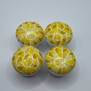 May include: Four round cabinet knobs with a mosaic design in shades of yellow and white. The knobs have a geometric pattern and are set against a white background. These decorative knobs can be used for furniture or other home decor projects.