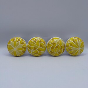 Peut inclure: Quatre boutons de meuble jaunes avec un motif de mosa&iuml;que. Les boutons sont ronds et pr&eacute;sentent un motif de formes g&eacute;om&eacute;triques jaunes et cr&egrave;me. Les boutons sont dispos&eacute;s en rang&eacute;e sur un fond blanc.