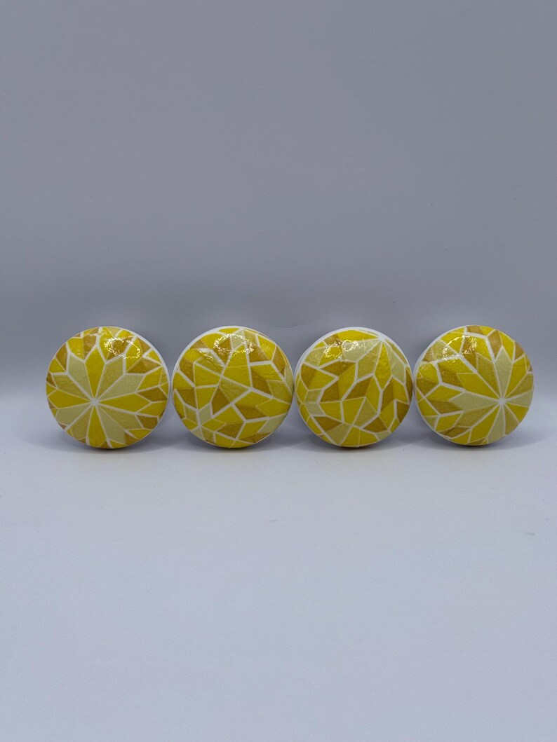 Peut inclure: Quatre boutons de meuble jaunes avec un motif de mosa&iuml;que. Les boutons sont ronds et pr&eacute;sentent un motif de formes g&eacute;om&eacute;triques jaunes et cr&egrave;me. Les boutons sont dispos&eacute;s en rang&eacute;e sur un fond blanc.