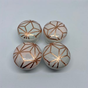 May include: Four white ceramic knobs with a geometric design in rose gold. The knobs are round and have a raised design.