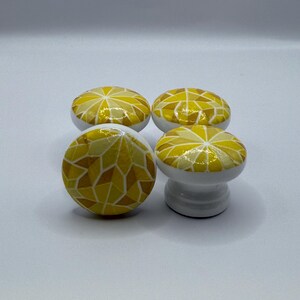 Peut inclure: Quatre boutons de meuble d&eacute;coratifs. Chaque bouton a une base blanche et un dessus en mosa&iuml;que dans des tons de jaune et d'or. Le motif de la mosa&iuml;que est compos&eacute; de formes g&eacute;om&eacute;triques irr&eacute;guli&egrave;res. Un bouton est sur un pi&eacute;destal.