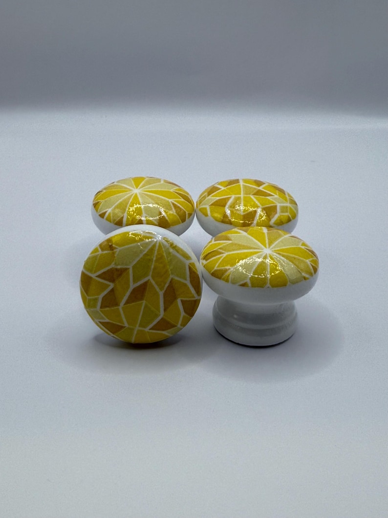 Peut inclure: Quatre boutons de meuble d&eacute;coratifs. Chaque bouton a une base blanche et un dessus en mosa&iuml;que dans des tons de jaune et d'or. Le motif de la mosa&iuml;que est compos&eacute; de formes g&eacute;om&eacute;triques irr&eacute;guli&egrave;res. Un bouton est sur un pi&eacute;destal.