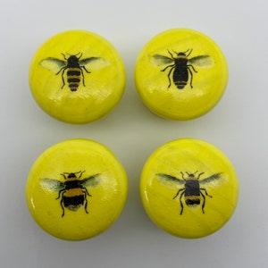 May include: Four yellow wooden knobs featuring a black and yellow bee design. The bees are painted onto the knobs and have black wings and a yellow body.
