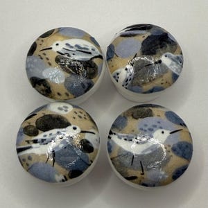 May include: Four circular cabinet knobs with a bird design. Each knob features a white bird with black details, set against a backdrop of tan, grey, and black abstract shapes. The knobs have a white base.