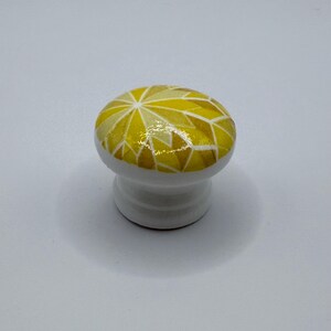 Peut inclure: Bouton de meuble en c&eacute;ramique blanche avec un motif g&eacute;om&eacute;trique dans les tons de jaune et d'or. Le bouton a un dessus arrondi et une base courte et cylindrique. Le motif g&eacute;om&eacute;trique est compos&eacute; de triangles et d'autres formes.