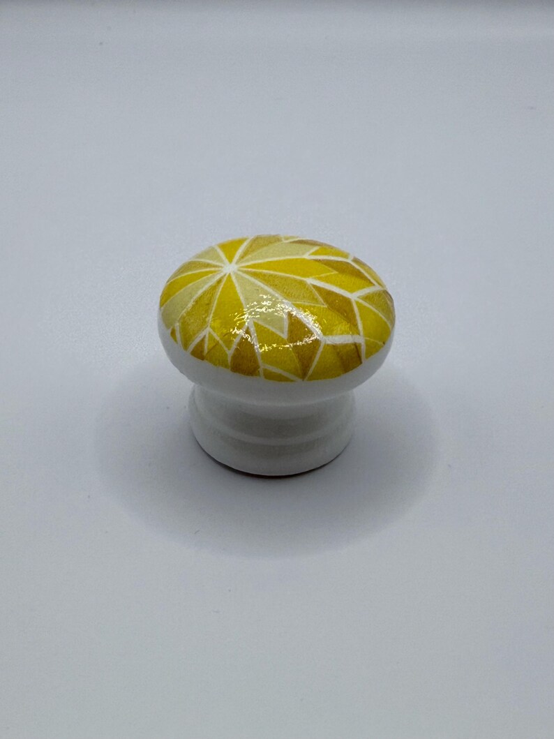 Peut inclure: Bouton de meuble en c&eacute;ramique blanche avec un motif g&eacute;om&eacute;trique dans les tons de jaune et d'or. Le bouton a un dessus arrondi et une base courte et cylindrique. Le motif g&eacute;om&eacute;trique est compos&eacute; de triangles et d'autres formes.