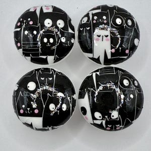 May include: Four circular cabinet knobs with a black and white cat design. Each knob has a glossy finish and a repeating pattern of stylised cat faces. The cats display different expressions against a black backdrop.