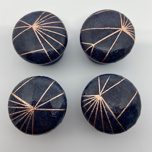 May include: Four round, dark blue cabinet knobs with a geometric design in rose gold. The knobs have a shiny, glossy finish.