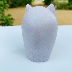 Może przedstawiać: Mała, jasnolilawa figurka ceramiczna w kształcie kota z zaokrąglonymi uszami. Figurka stoi na białym tle.